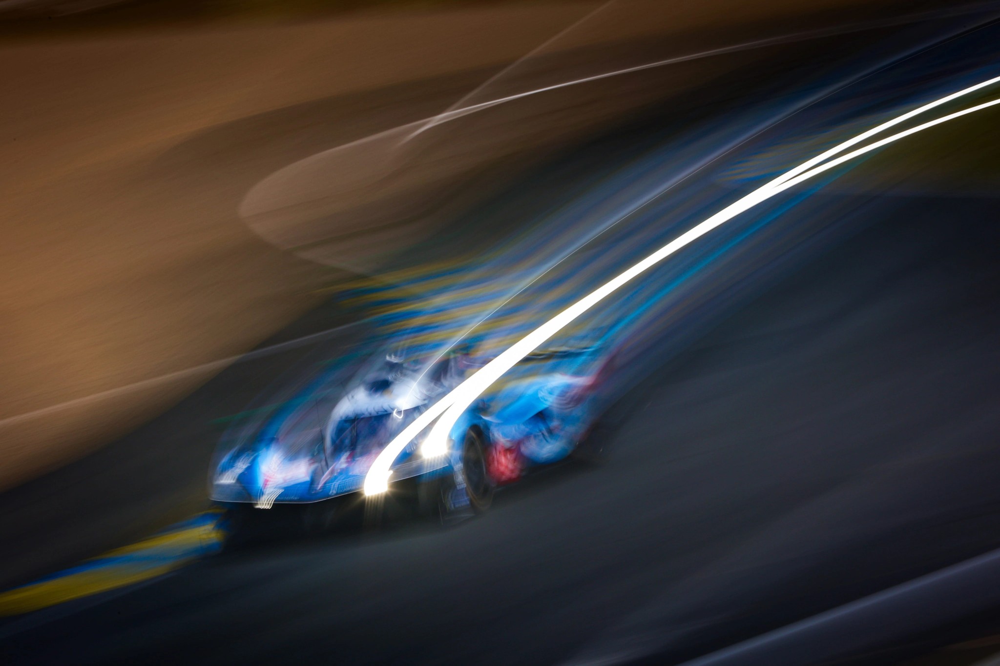 Une voiture de course bleue roule de nuit sur le circuit, ses phares traçant de longues lignes lumineuses