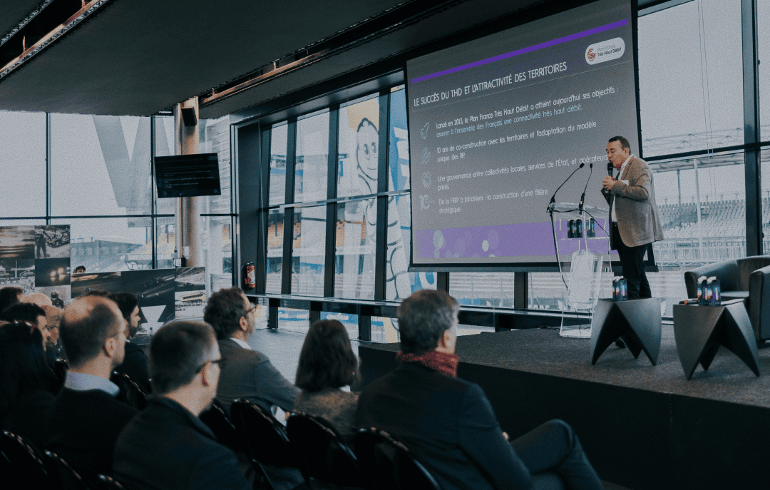 Conférencier sur scène devant un écran de présentation et un public assis dans une salle vitrée