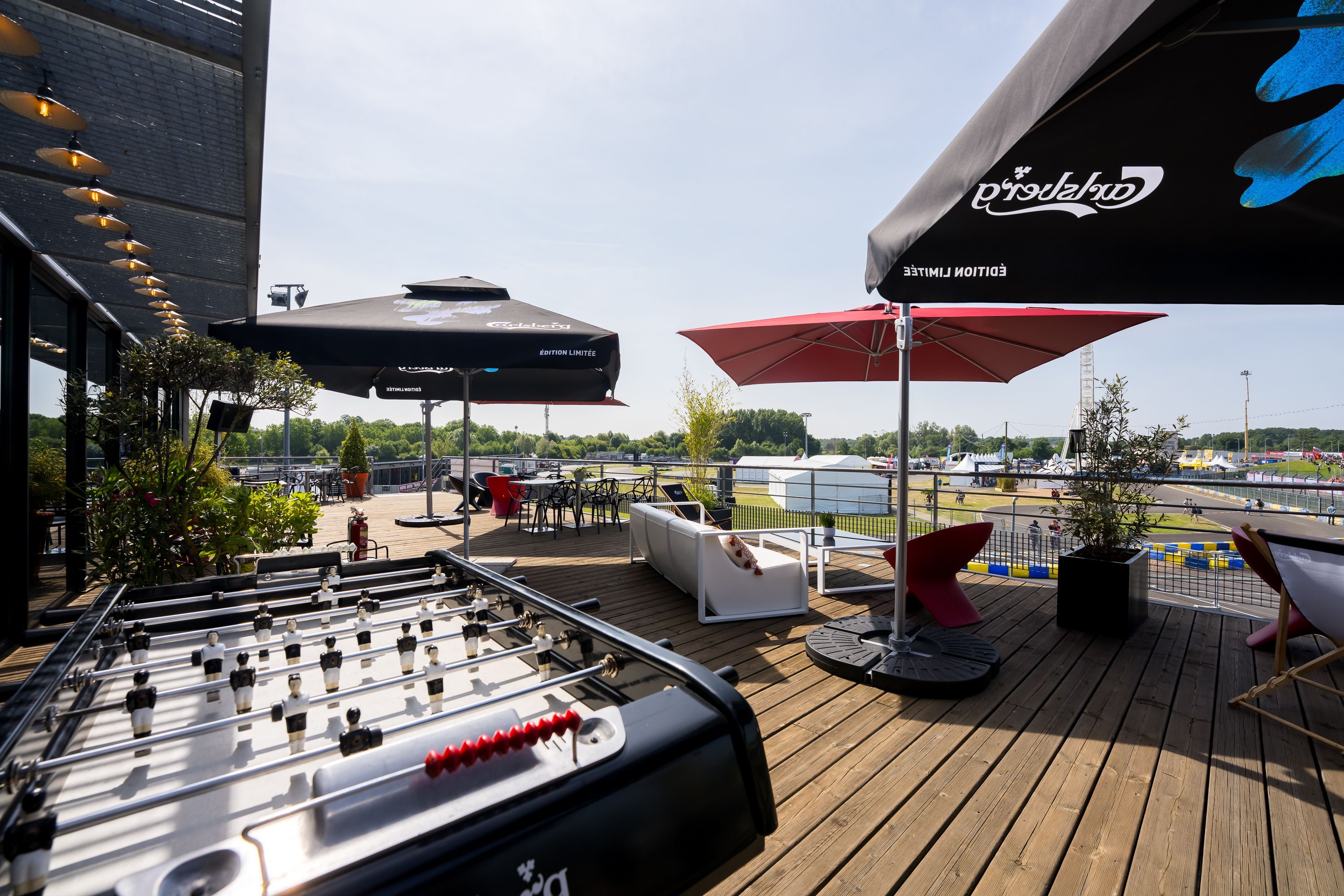 Terrasse avec baby-foot, parasols, tables et fauteuils donnant sur le circuit des 24 Heures du Mans