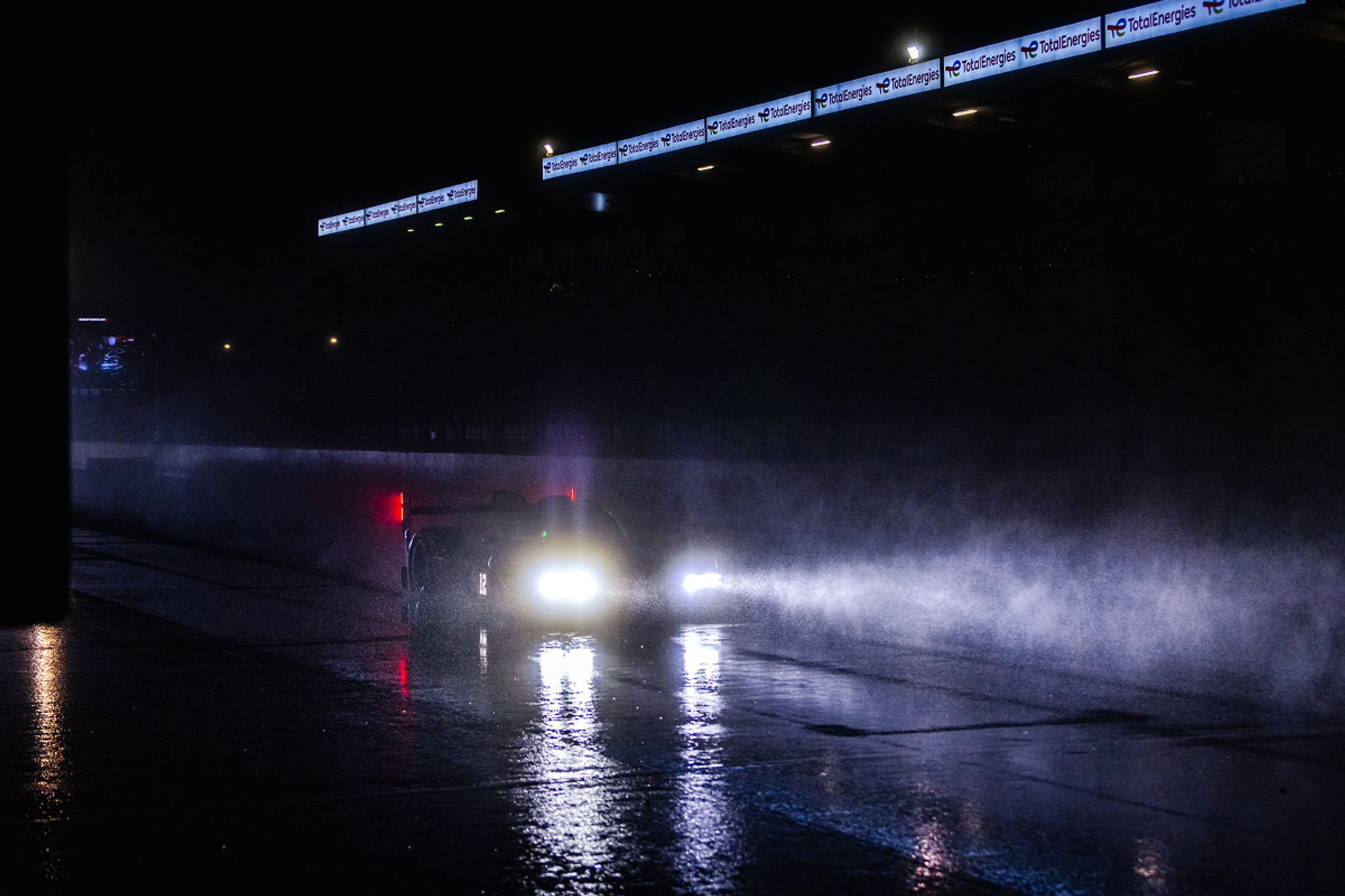 Voiture de course roulant de nuit sous la pluie, phares allumés et gerbes d’eau sur la piste