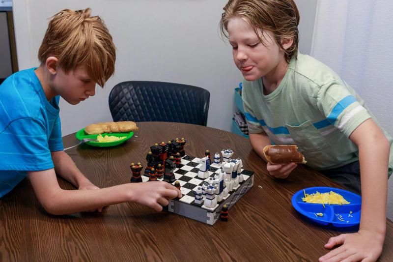 Two boys play chess with Lego pieces. One boy moves a piece while the other holds a donut and licks his lips..
