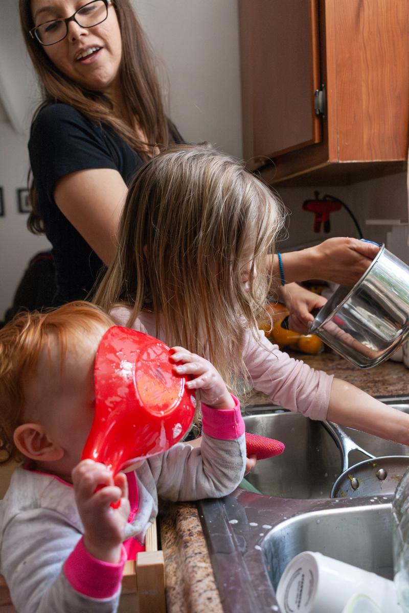 A woman oversees two young children at a kitchen sink; one washes a pot, and the other plays with a red bowl.