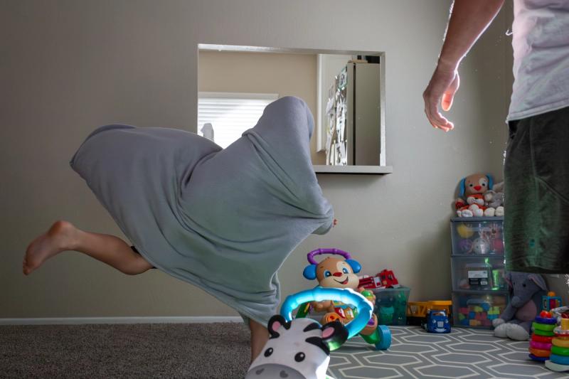 A child covered in a gray blanket plays in a toy-filled room, with an adult partially visible on the right.