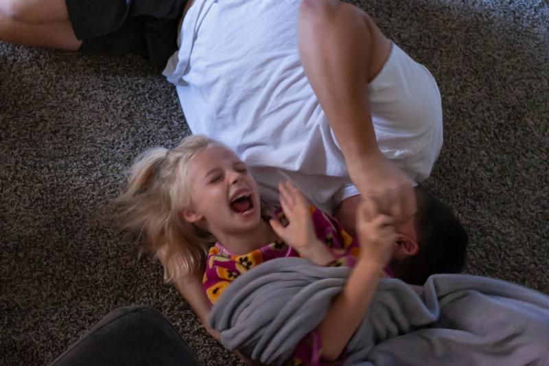 A blonde child laughs joyfully while playing on a carpet with a parent.