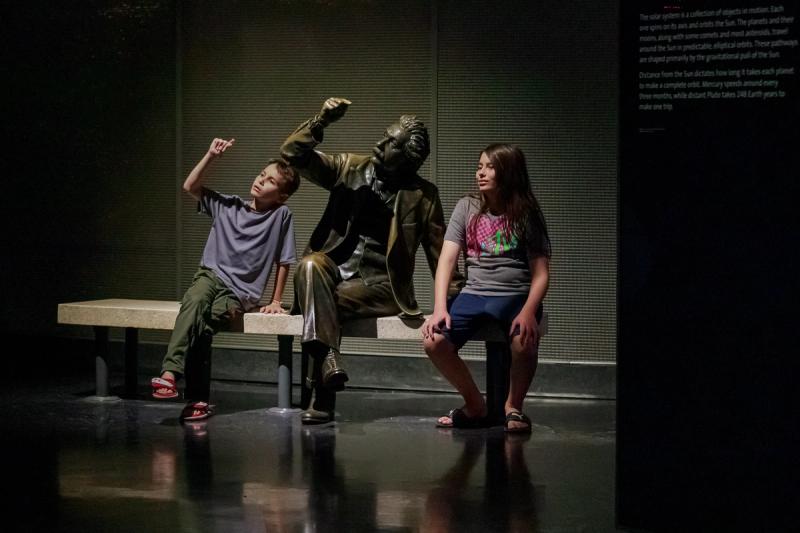 A boy and a bronze statue sit on a bench, both pointing upwards, while another boy sits next to them on the edge of the bench.