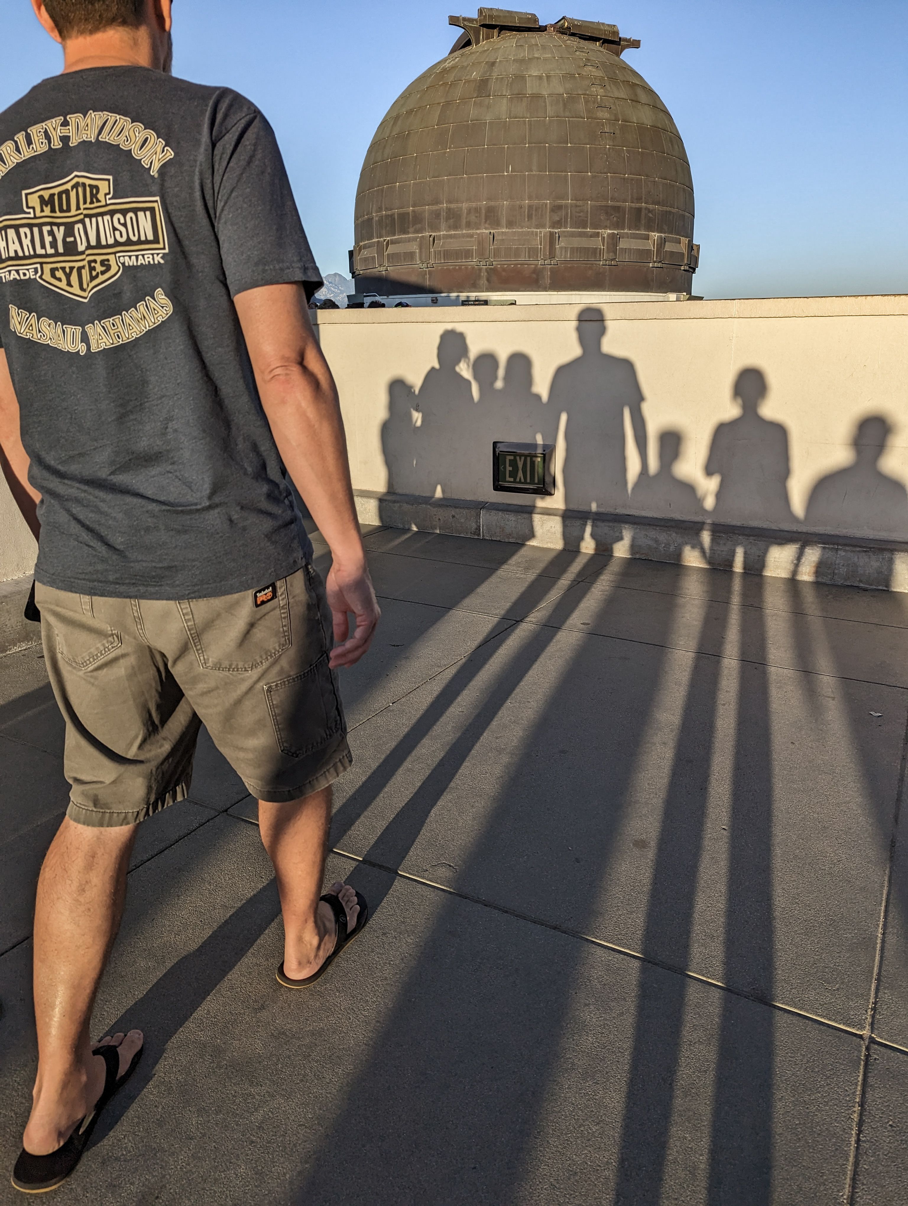 Man walking across rooftop terrace at sunset with long shadows of people cast toward an observatory dome.