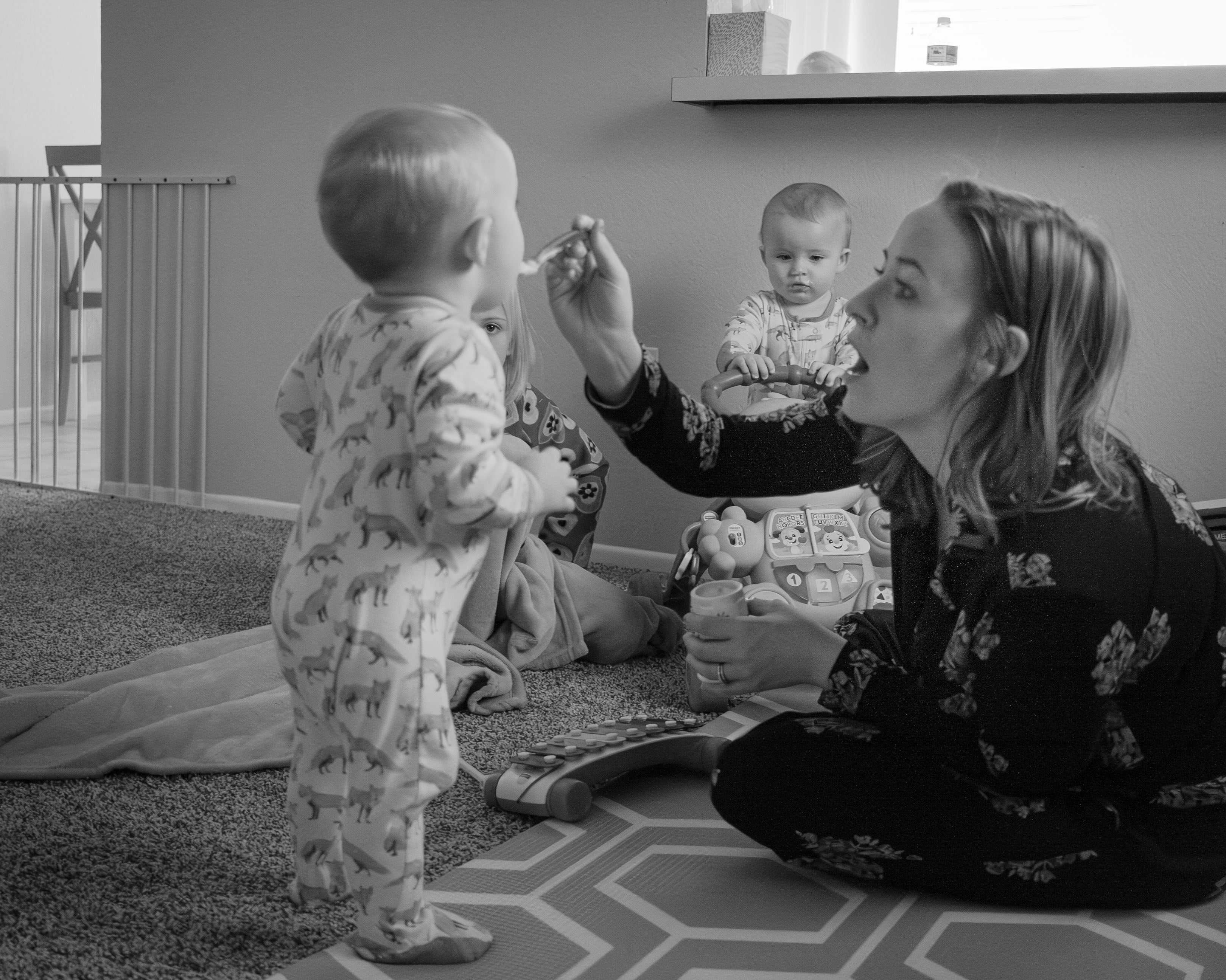 A mom feeding her toddler on the floor in a playroom with two other children watching