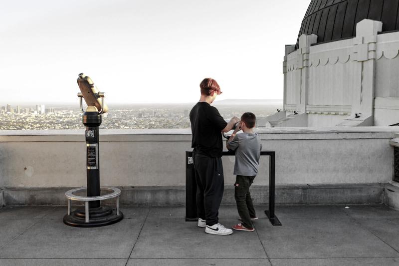 A teenager and a child look out over a city from an observation deck, with a public telescope and an observatory dome.