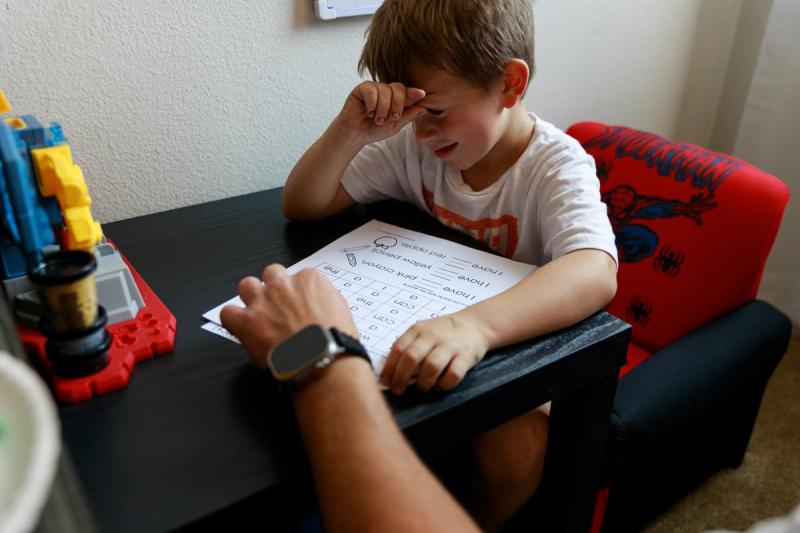 A young boy pauses, hand on head, while working on homework at a desk with an adult.