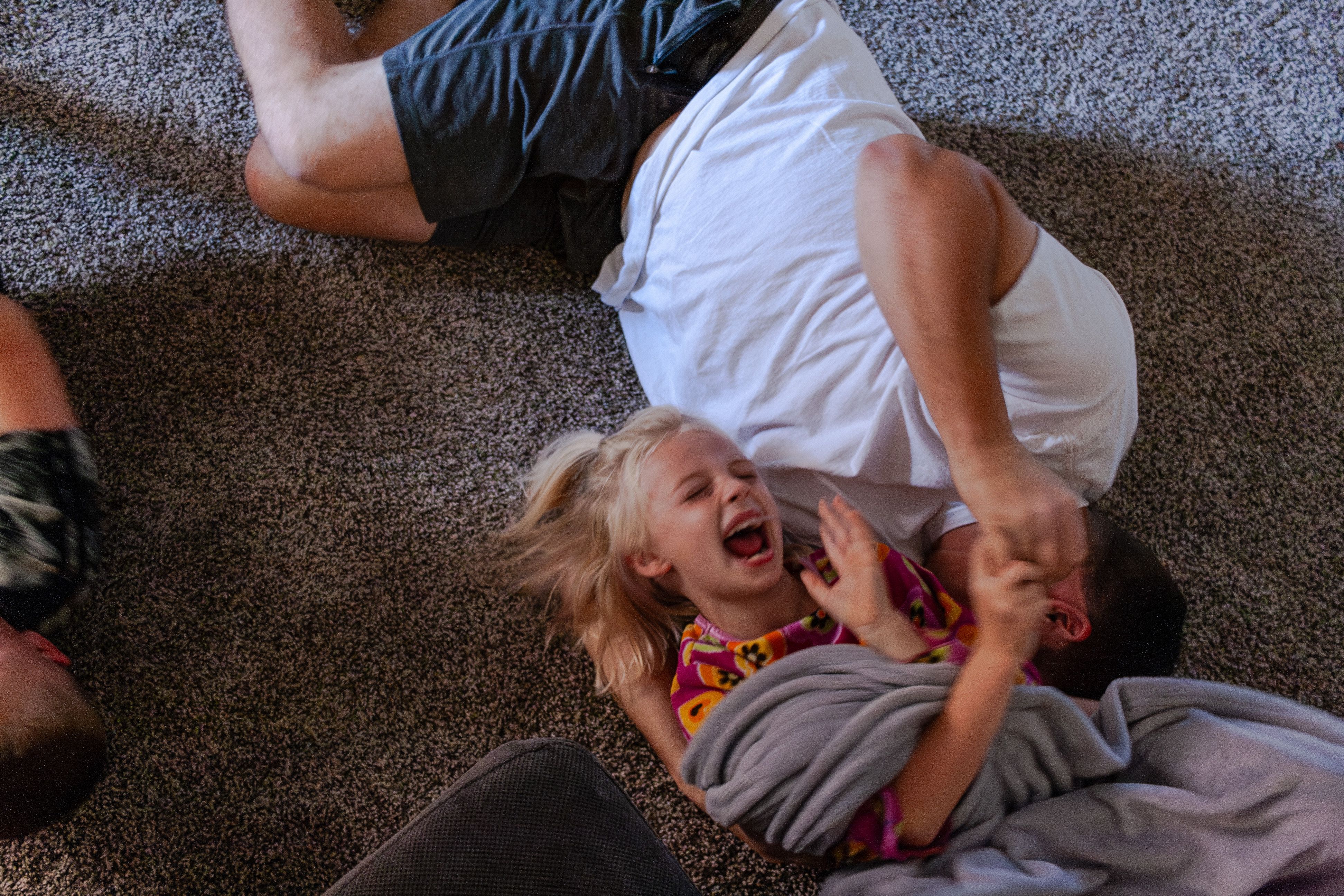 A young girl laughs joyfully while playfully wrestling on a carpeted floor with an adult, wrapped in a soft gray blanket. The candid moment captures energetic, carefree fun at home.