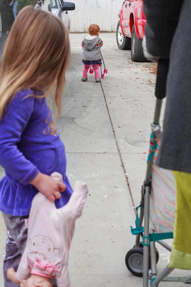 An older child holding a doll looks at a younger child pushing a toy stroller away on a concrete path.