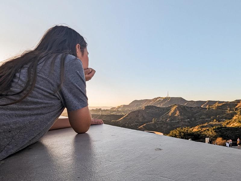Child from behind leaning on a ledge, gazing at the Hollywood Sign amidst golden-lit hills.