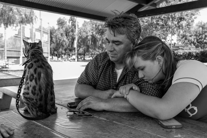 A father and daughter, the daughter resting her head on his shoulder, sit at a picnic table next to a leashed spotted cat. Black and white image.
