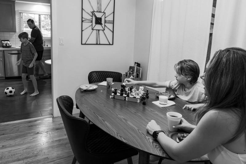 Black and white photo of a family indoors: a woman and boy sit at a dining table, while another boy plays with a soccer ball.