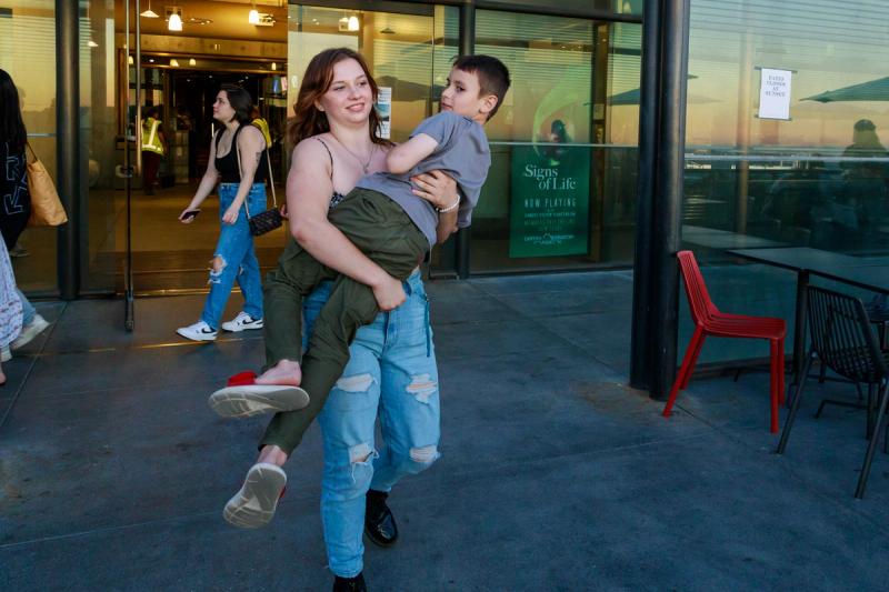 A smiling woman carries a young boy in her arms, walking past glass-front buildings with outdoor seating.