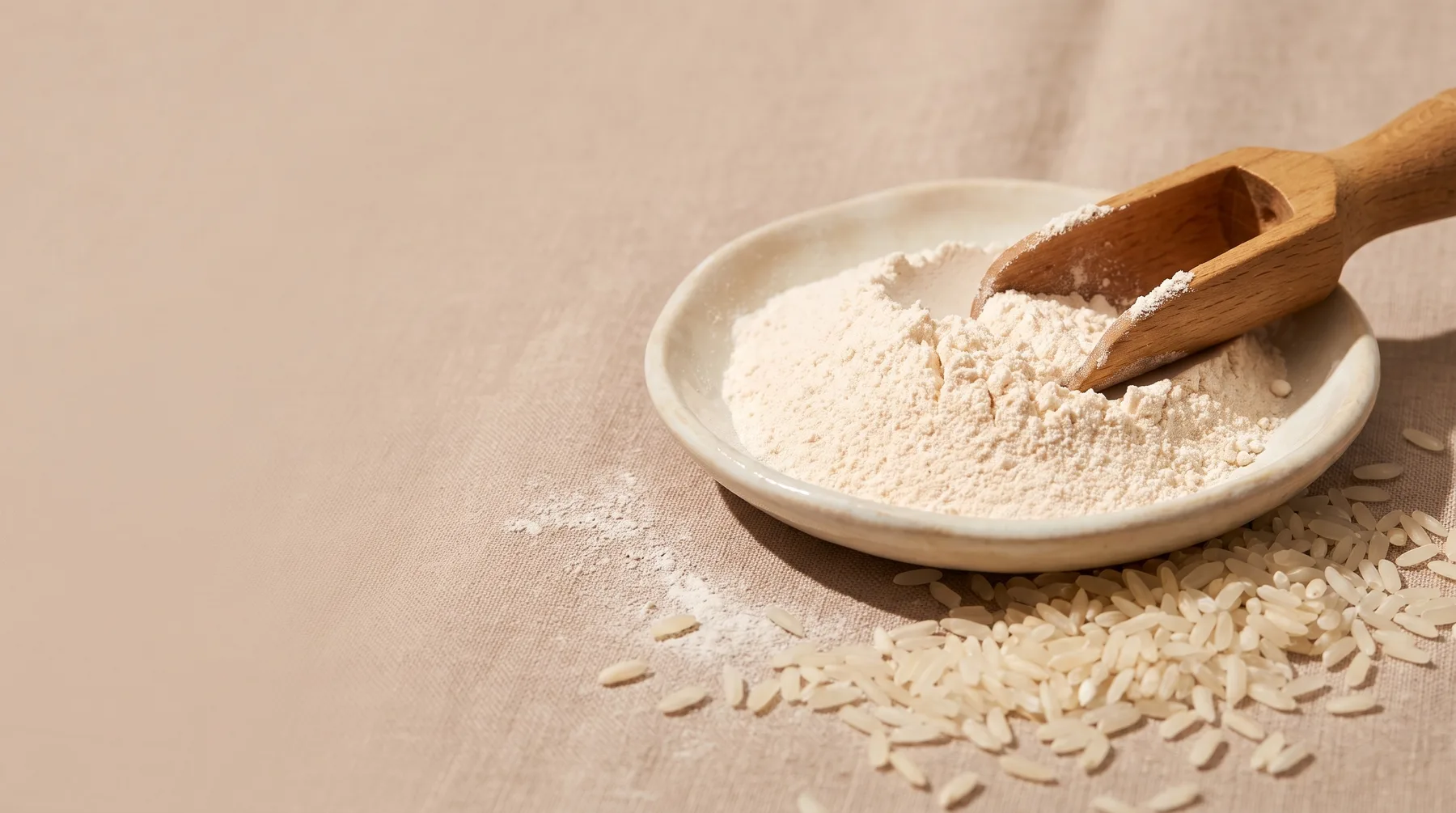 Kojic acid — pale kojic acid powder in a ceramic dish with a wooden scoop and scattered rice grains on warm linen