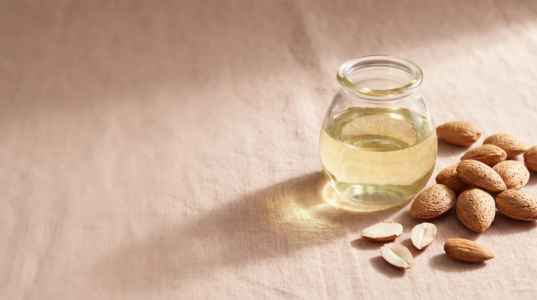 Almond oil — a glass vessel of sweet almond oil beside whole and split almonds on warm linen