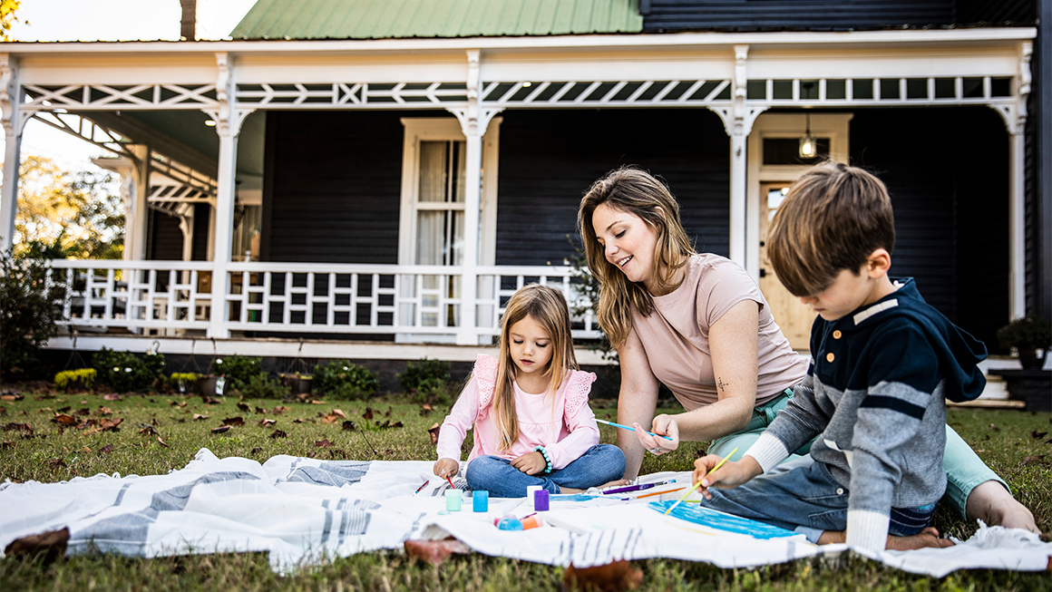 A family sits in front of a house on a blanket in the grass.