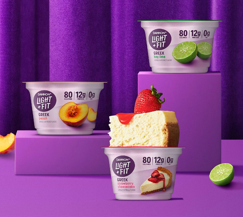 3 light and fit greek yogurt packages on purple pedastals against a purple curtain with fruits scattered around like a peach, strawberry and lime  - beardwood