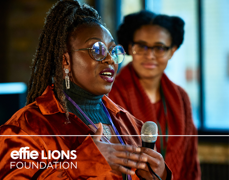 photo of a woman speaking holding a microphone with a woman watching behind her and an effie lions foundation logo in the lower left corner - beardwood
