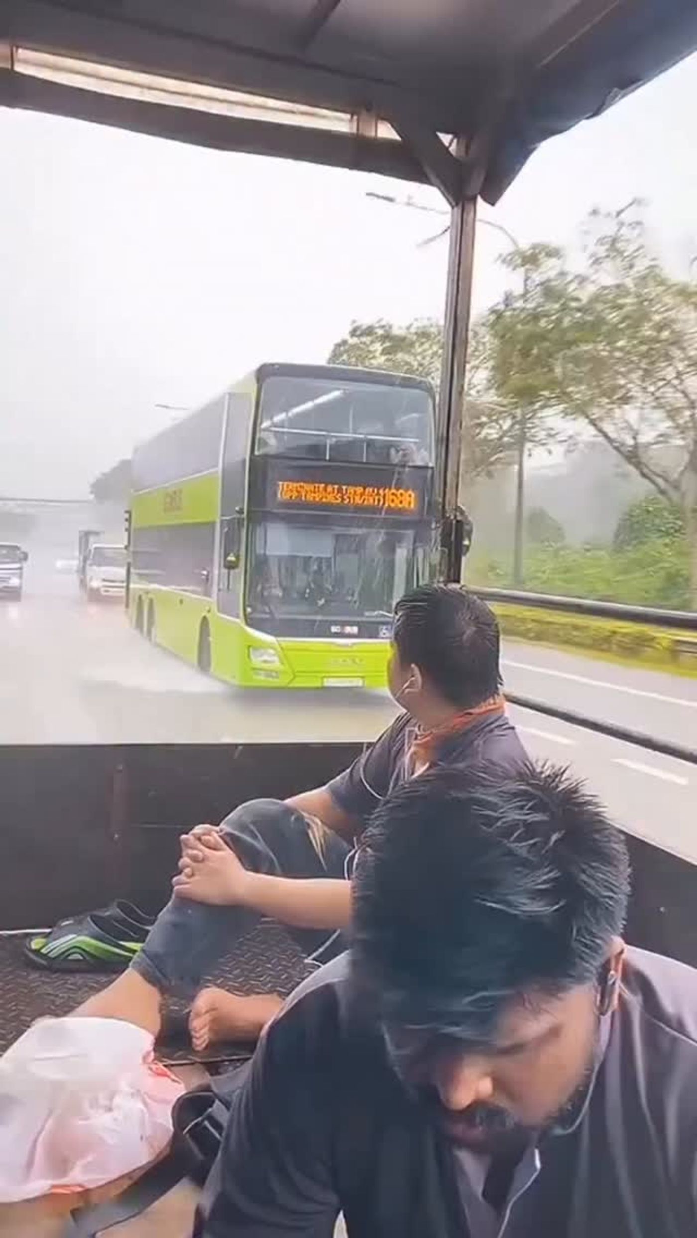 Passengers ride in open-backed vehicle as bus passes during heavy rain
