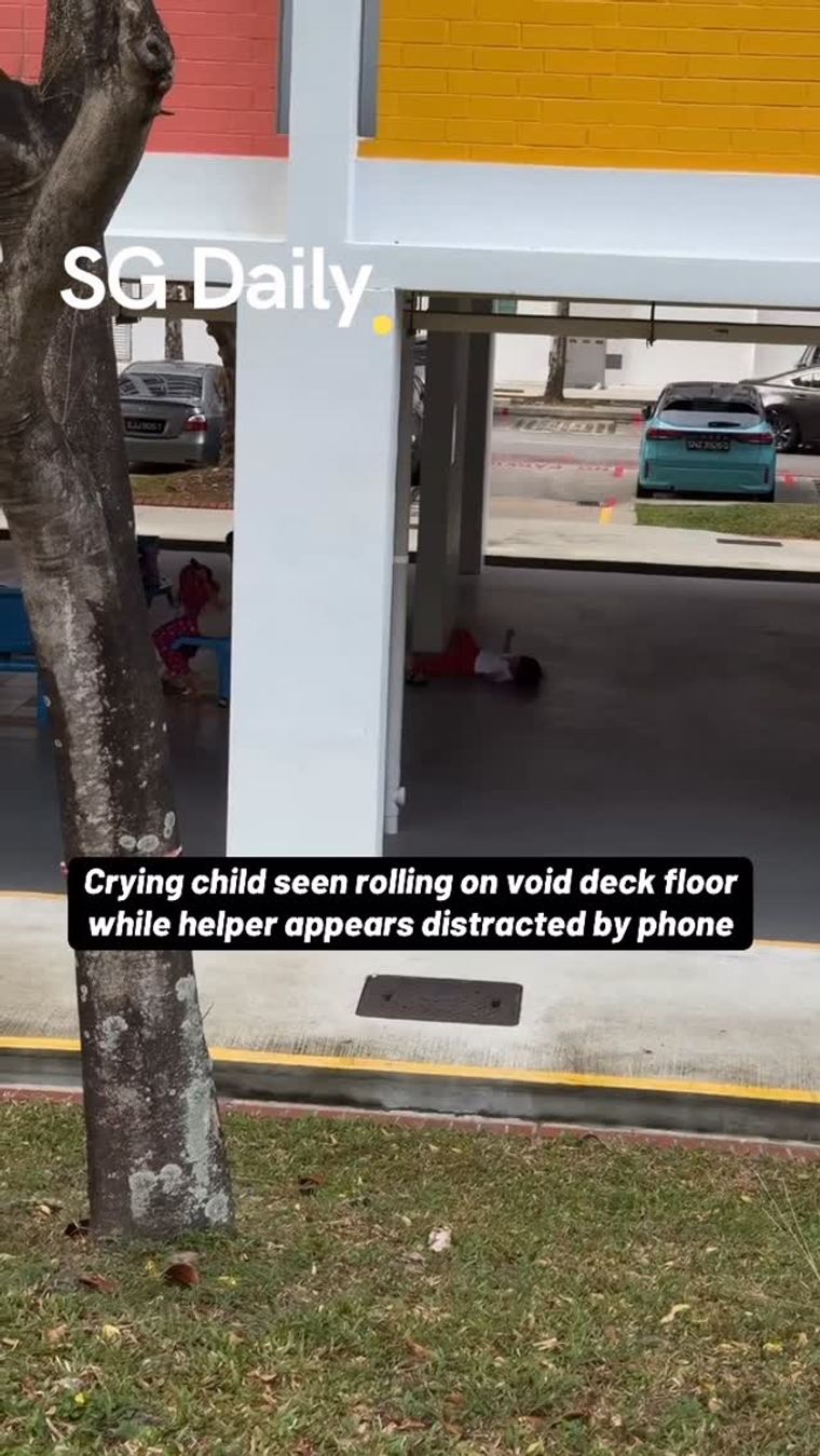 Crying child seen rolling on void deck floor while helper appears distracted by phone
