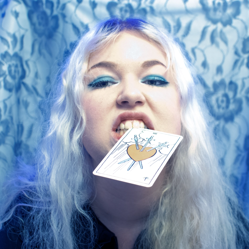 Blonde woman with teal eyeliner gripping the pierced-heart tarot card between her teeth, blue lace behind