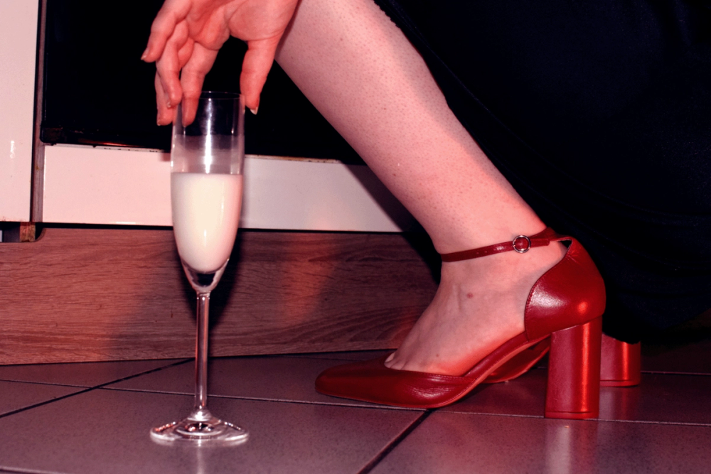 Hand lowering a champagne flute of milky liquid to the floor beside red block-heel sandals and a black hem