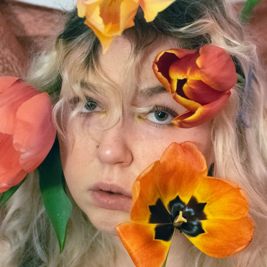 Close-up self-portrait, four tulips in coral and orange framing the face, green eyes fixed just past the lens