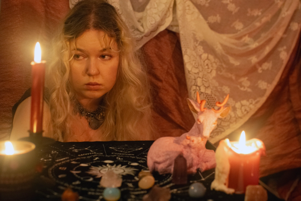 Blonde figure staring sideways behind a candlelit table of crystals, rust drapery and lace filling the room