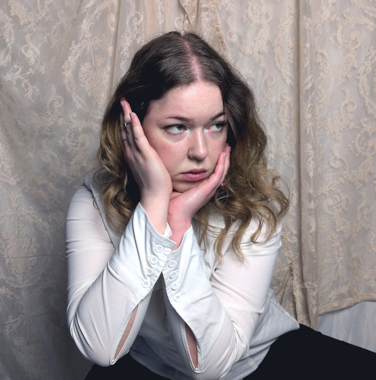 Sarah Massarczyk seated in a white blouse, both hands cradling her face, before a pale damask curtain