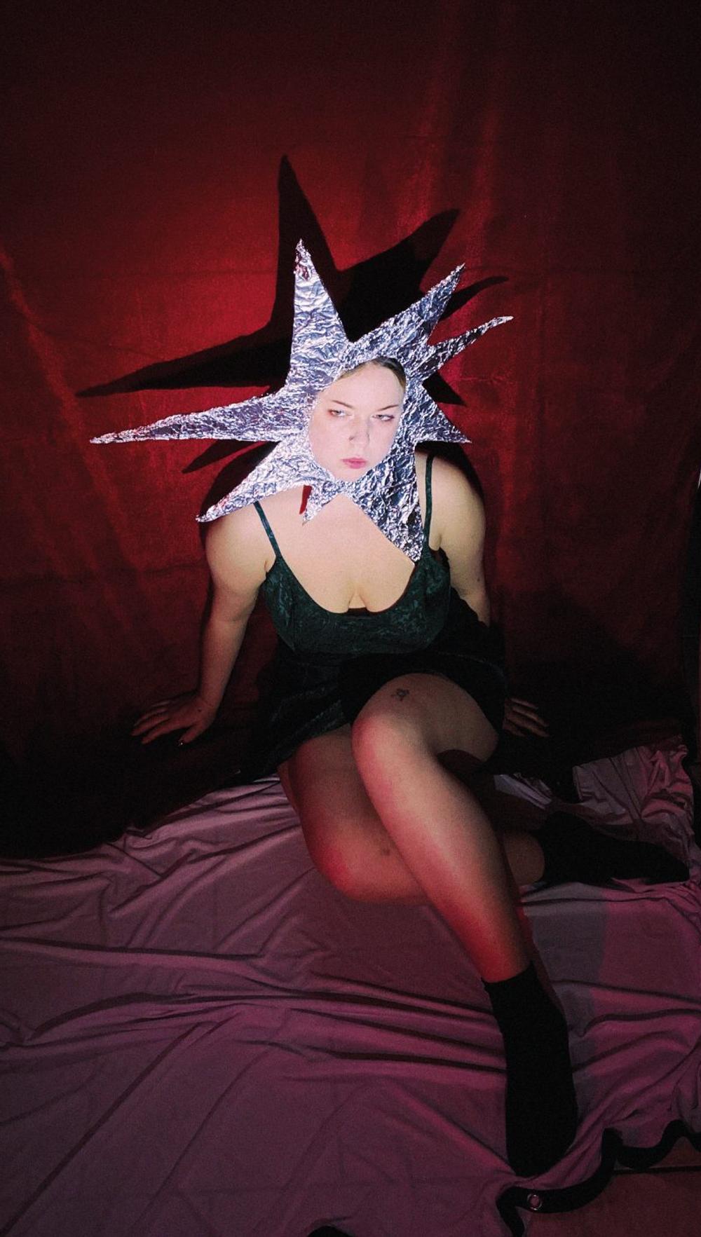 Silver foil star headpiece covering the face, green slip dress, seated against a heavy red backdrop