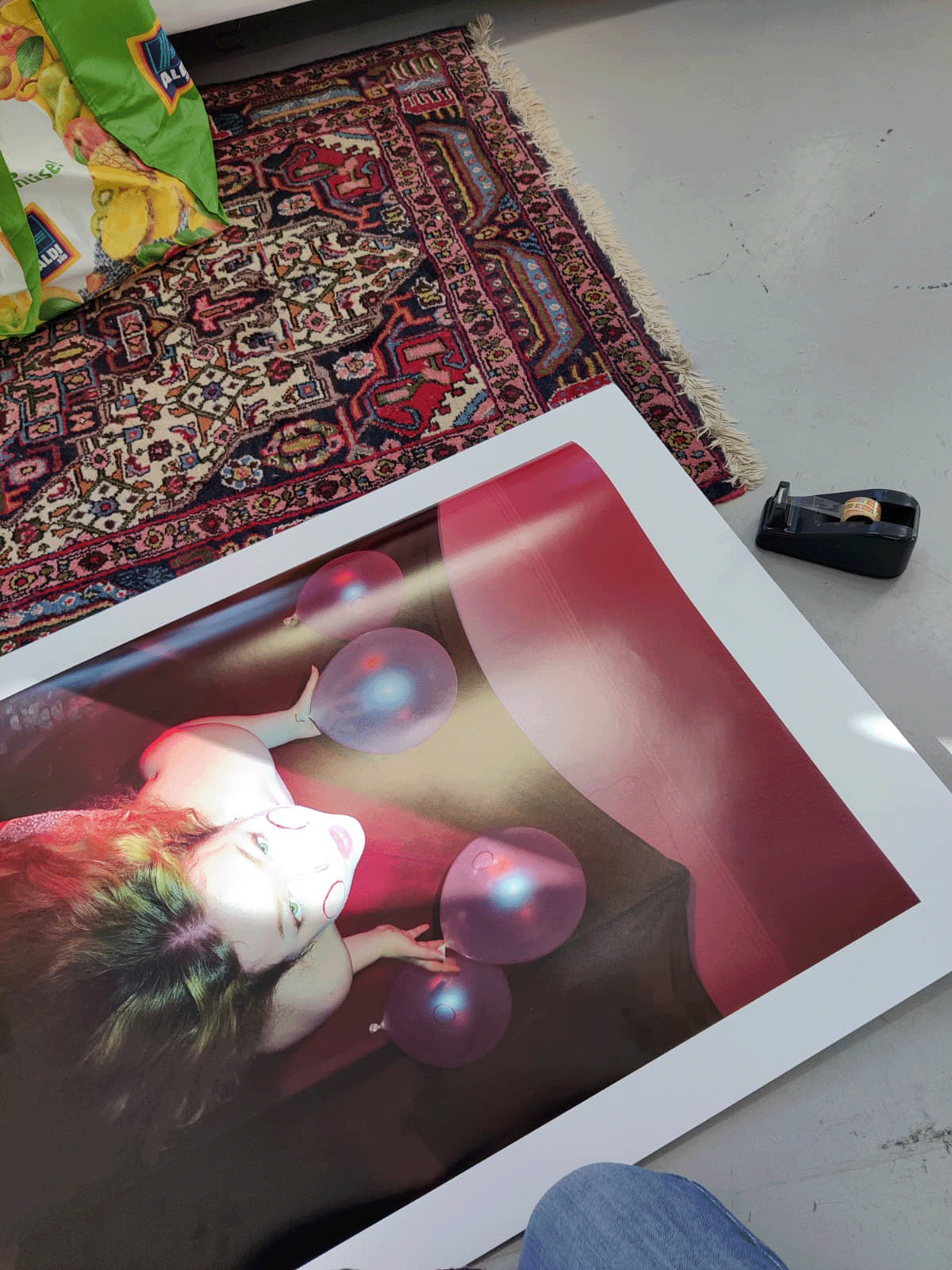 Large glossy print lying on a concrete floor beside a Persian rug, showing a figure with violet balloons