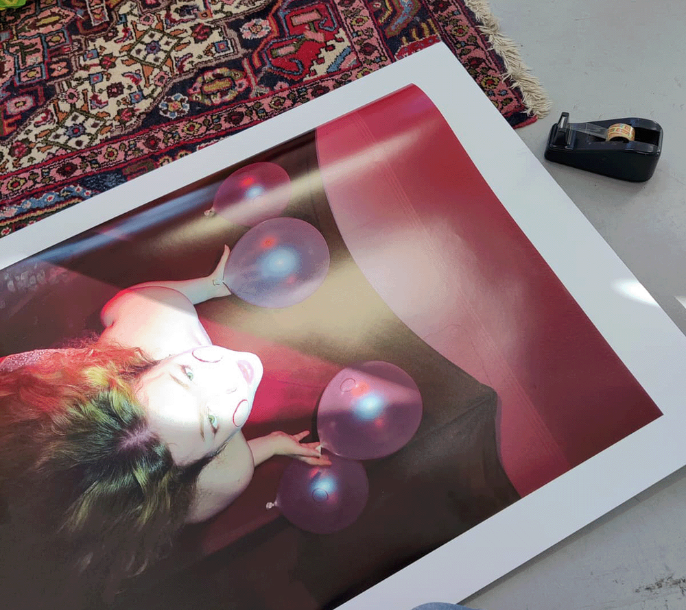 Large glossy print lying on a concrete floor beside a Persian rug, showing a figure with violet balloons