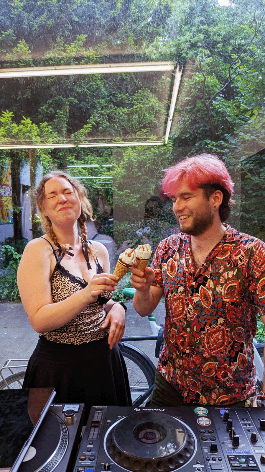 Two DJs behind a Pioneer setup outdoors, holding soft-serve cones, ivy and neon tubes filling the summer backdrop