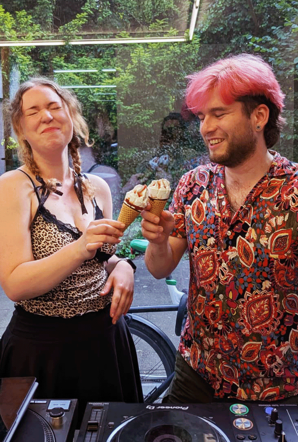 Two DJs behind a Pioneer setup outdoors, holding soft-serve cones, ivy and neon tubes filling the summer backdrop