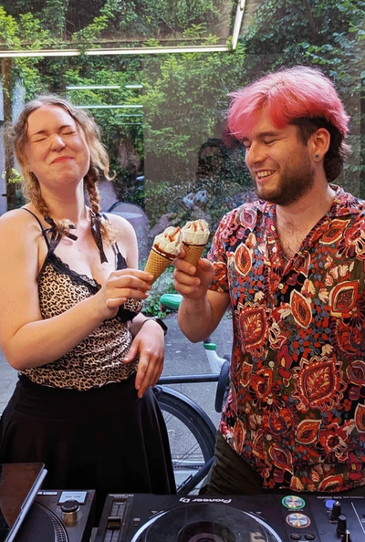 Two DJs behind a Pioneer setup outdoors, holding soft-serve cones, ivy and neon tubes filling the summer backdrop
