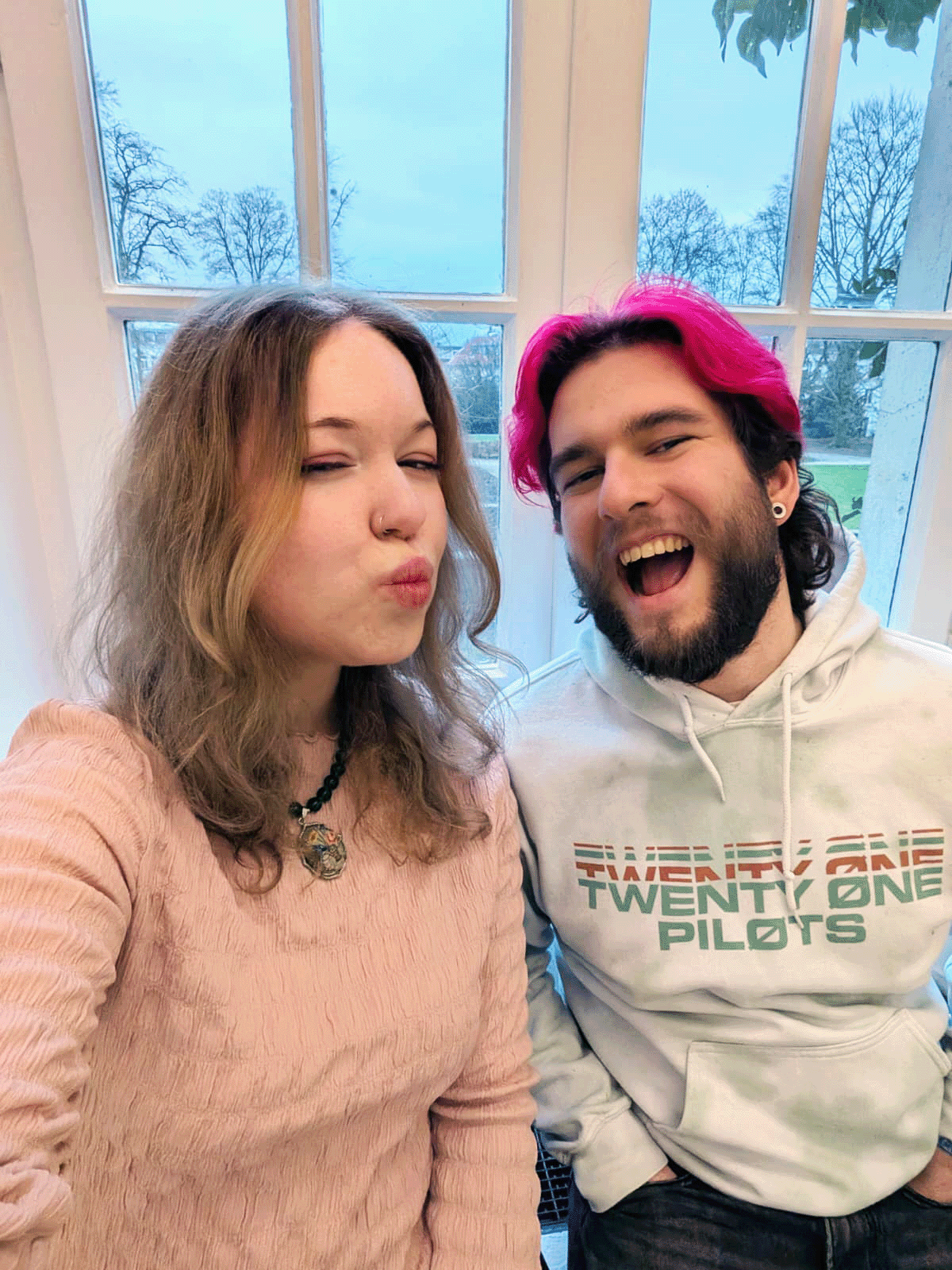Selfie of two hosts at a tall window, one pouting in a pink knit, the other pink-haired in a Twenty One Pilots hoodie