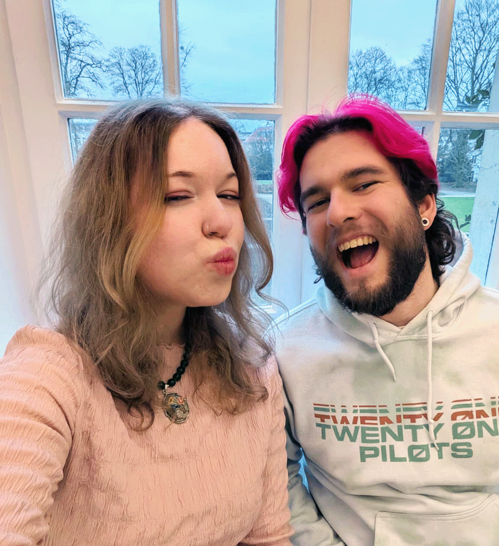 Selfie of two hosts at a tall window, one pouting in a pink knit, the other pink-haired in a Twenty One Pilots hoodie