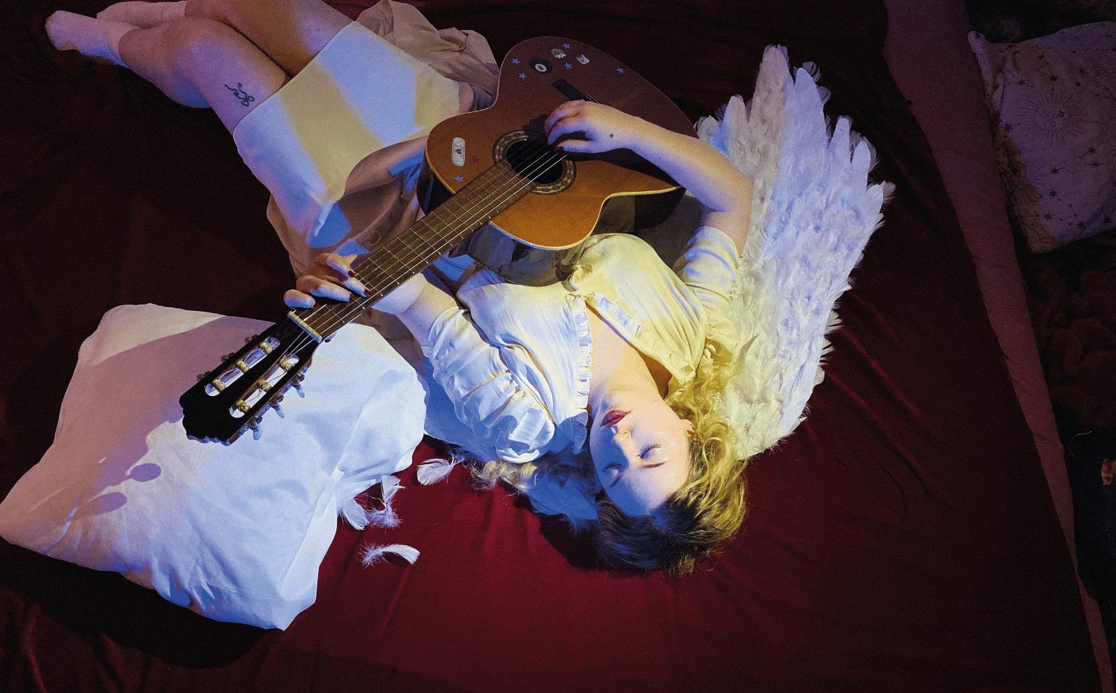 Seen from above, lying on deep red bedding in white angel wings with an acoustic guitar across the body
