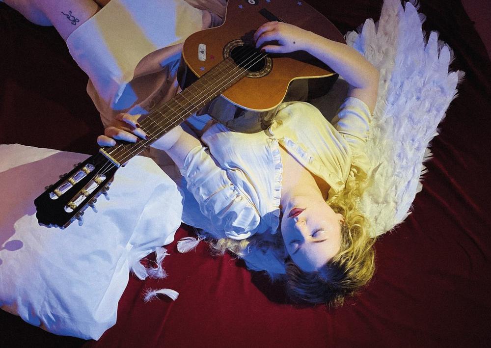 Seen from above, lying on deep red bedding in white angel wings with an acoustic guitar across the body