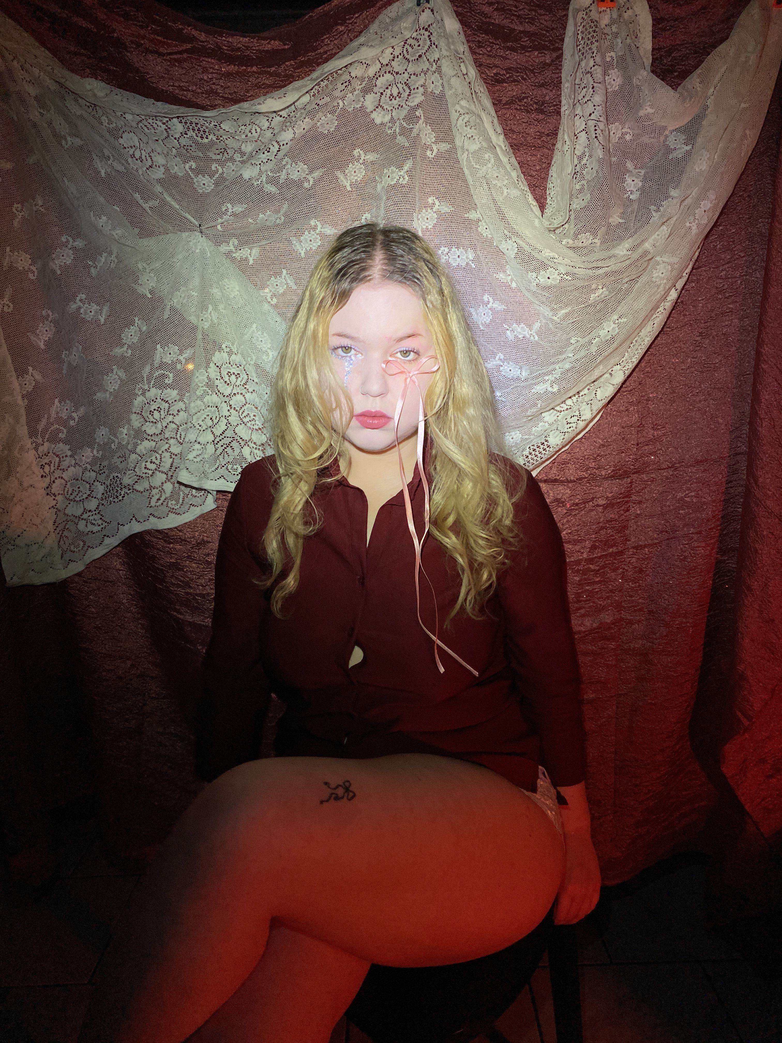 Seated on a stool in a dark red shirt, a pink ribbon trailing from the eye, ivory lace draped behind