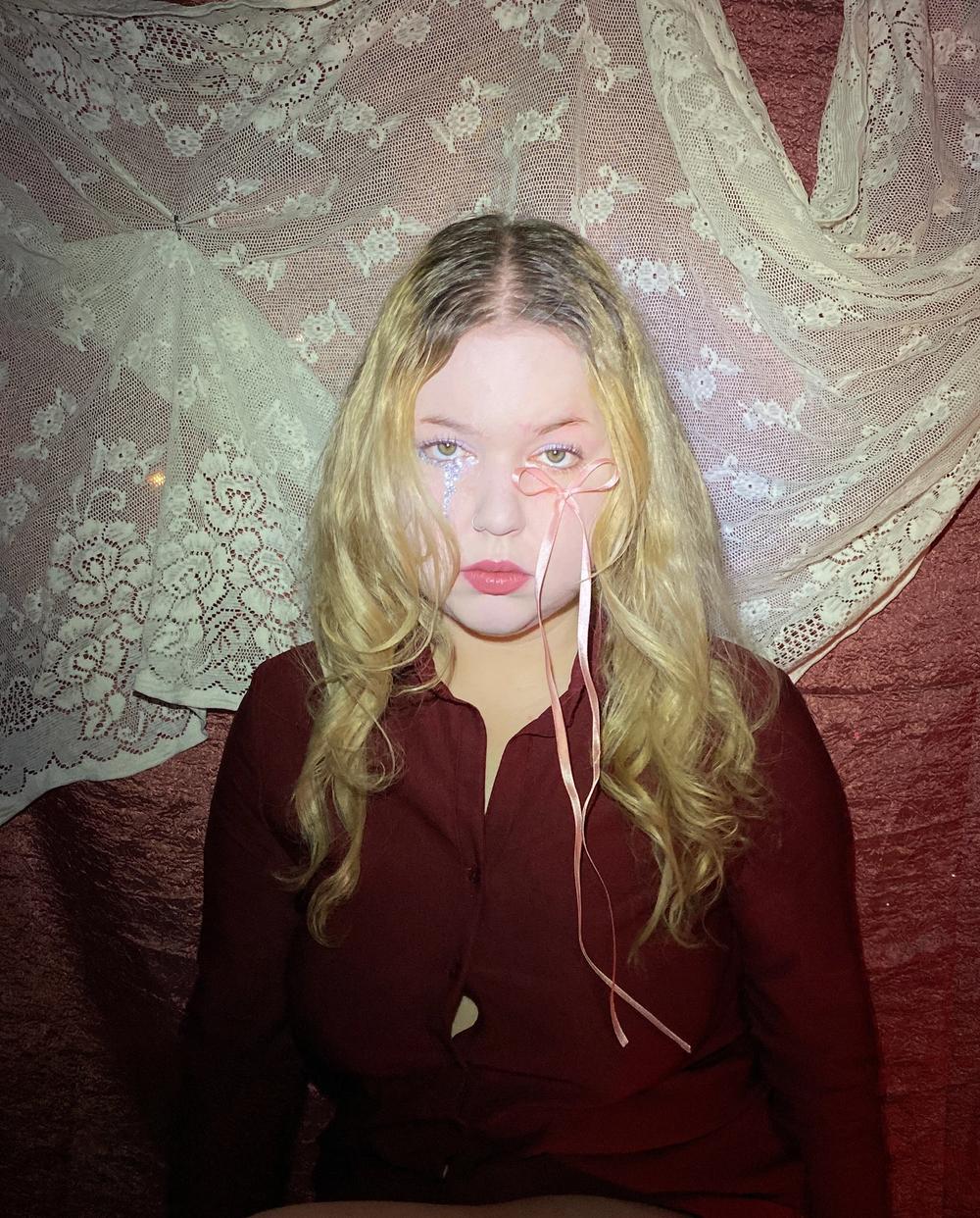 Seated on a stool in a dark red shirt, a pink ribbon trailing from the eye, ivory lace draped behind