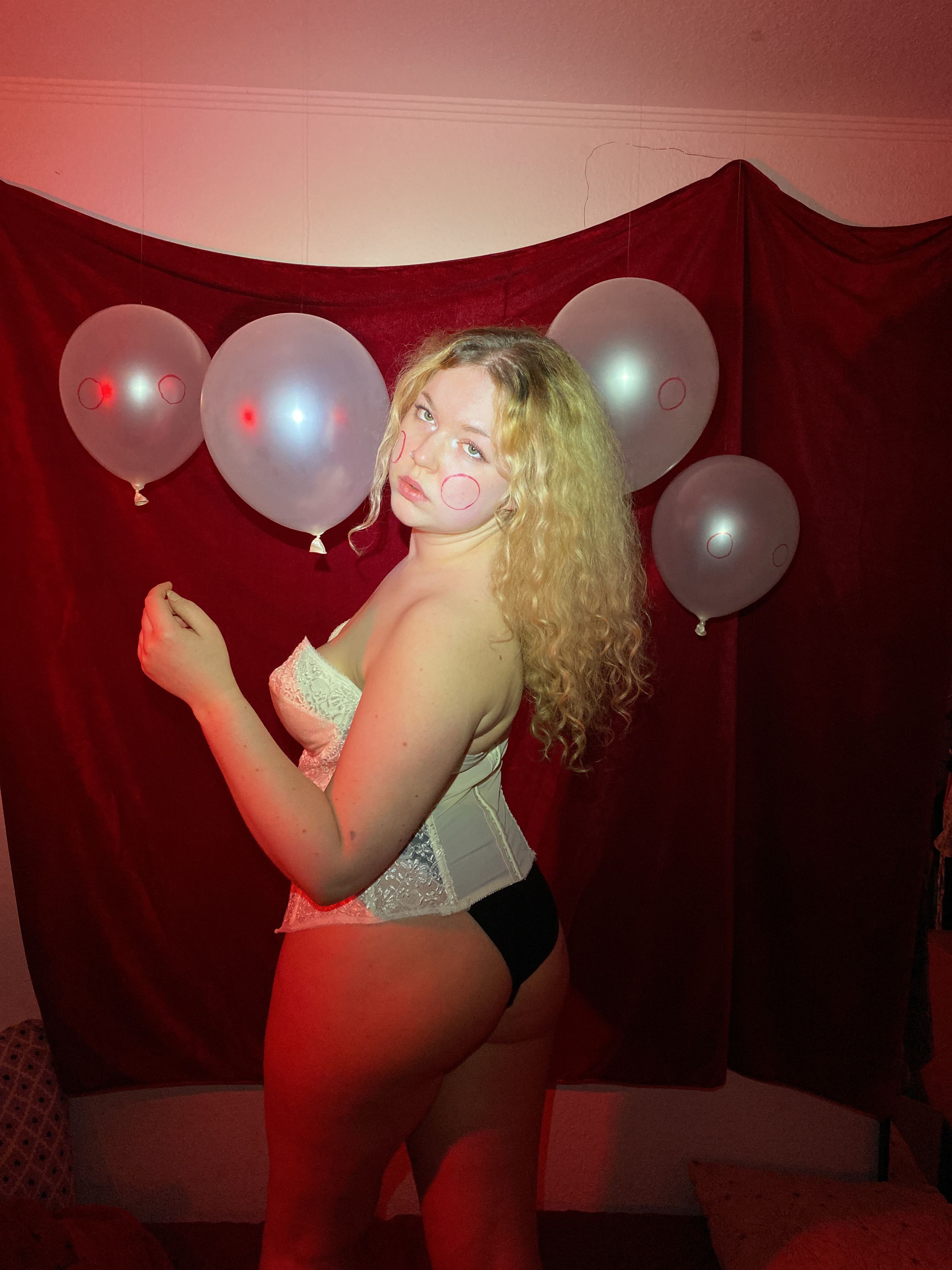 Red-lit self-portrait in a white corset, pale balloons marked with blush circles pinned to a crimson curtain