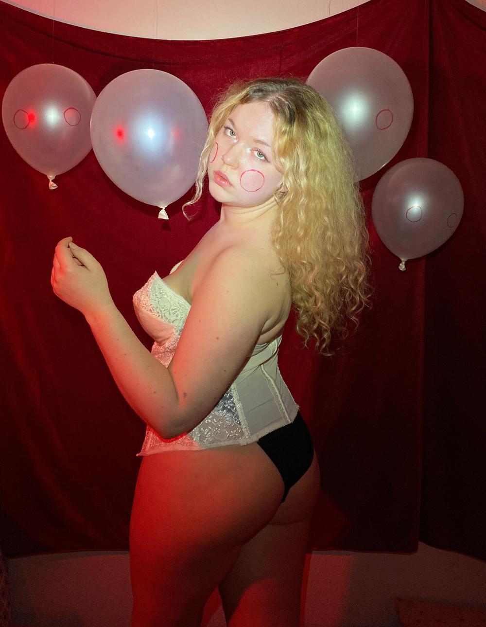 Red-lit self-portrait in a white corset, pale balloons marked with blush circles pinned to a crimson curtain