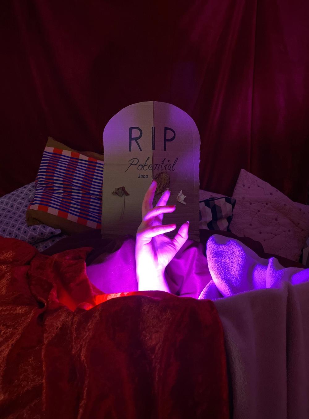 A hand rising from red bedding to hold a cardboard gravestone lettered RIP Potential in violet light