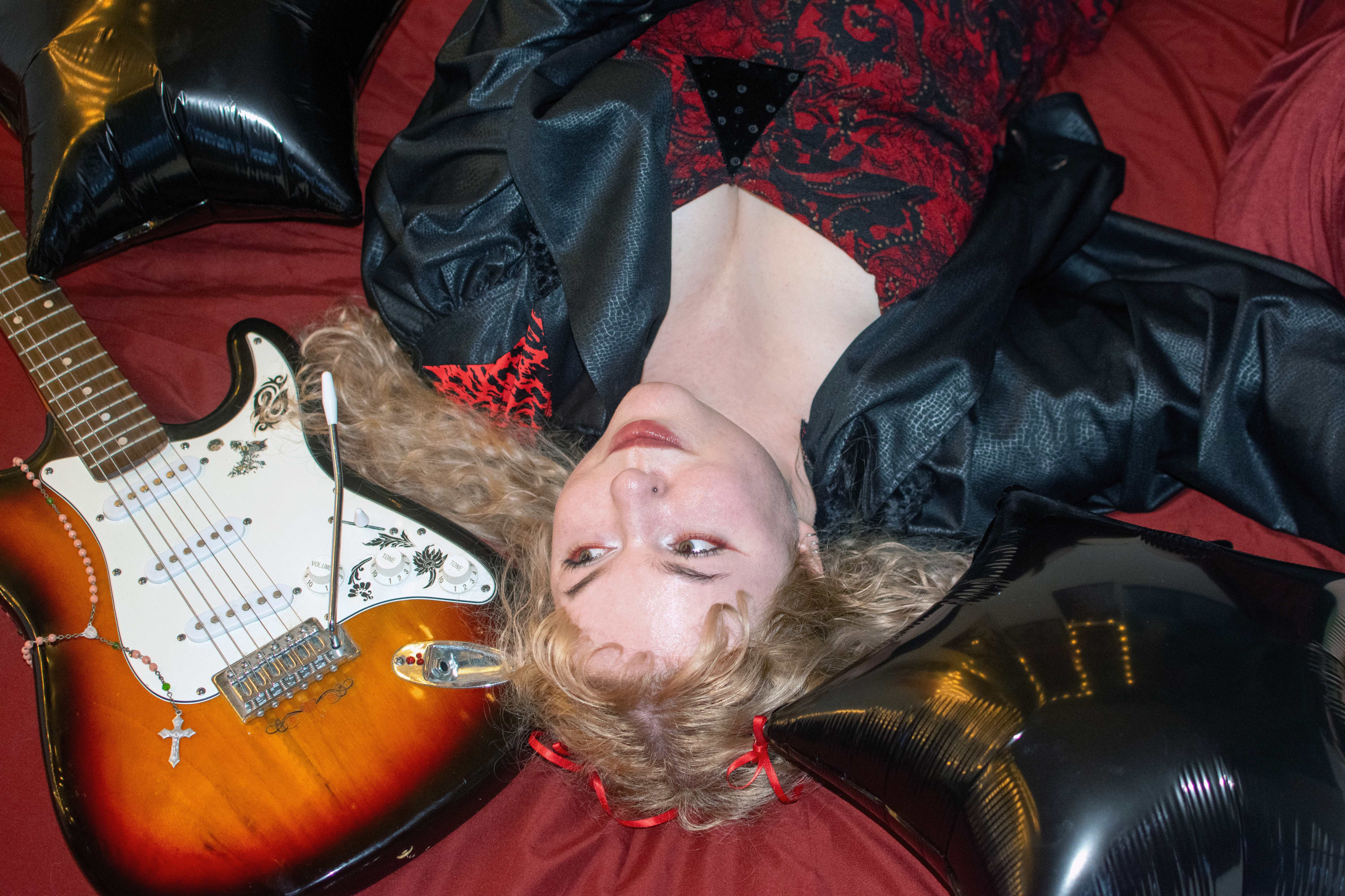 Sarah Massarczyk lying head-back on red bedding beside a sunburst electric guitar and black foil balloons