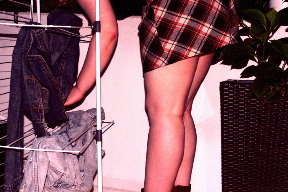 Bare legs in a red tartan mini skirt facing a drying rack hung with denim jackets, wall lit pink