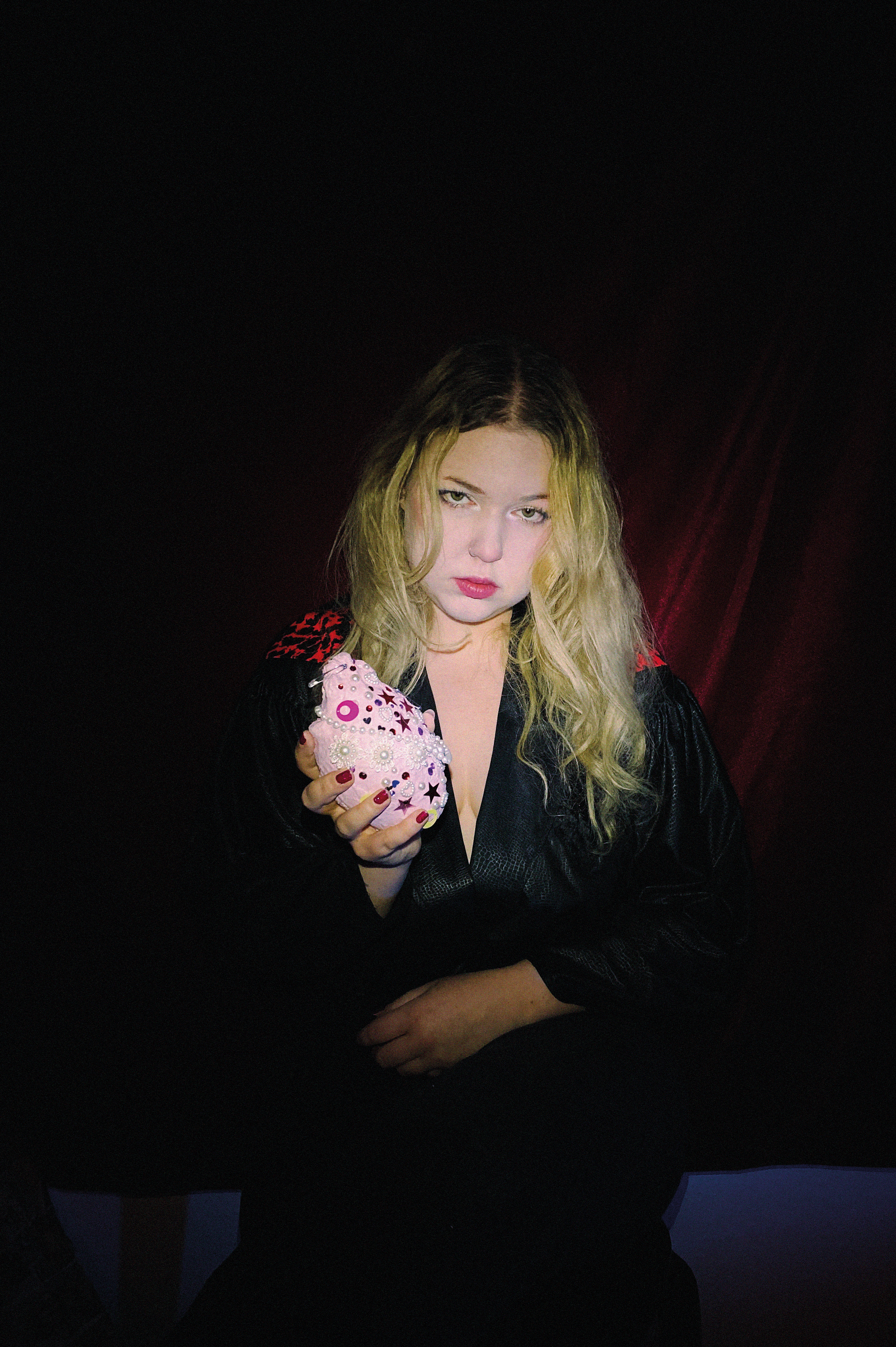 Low-key portrait in a black robe, holding out a soft pink heart studded with pearls, beads and stars