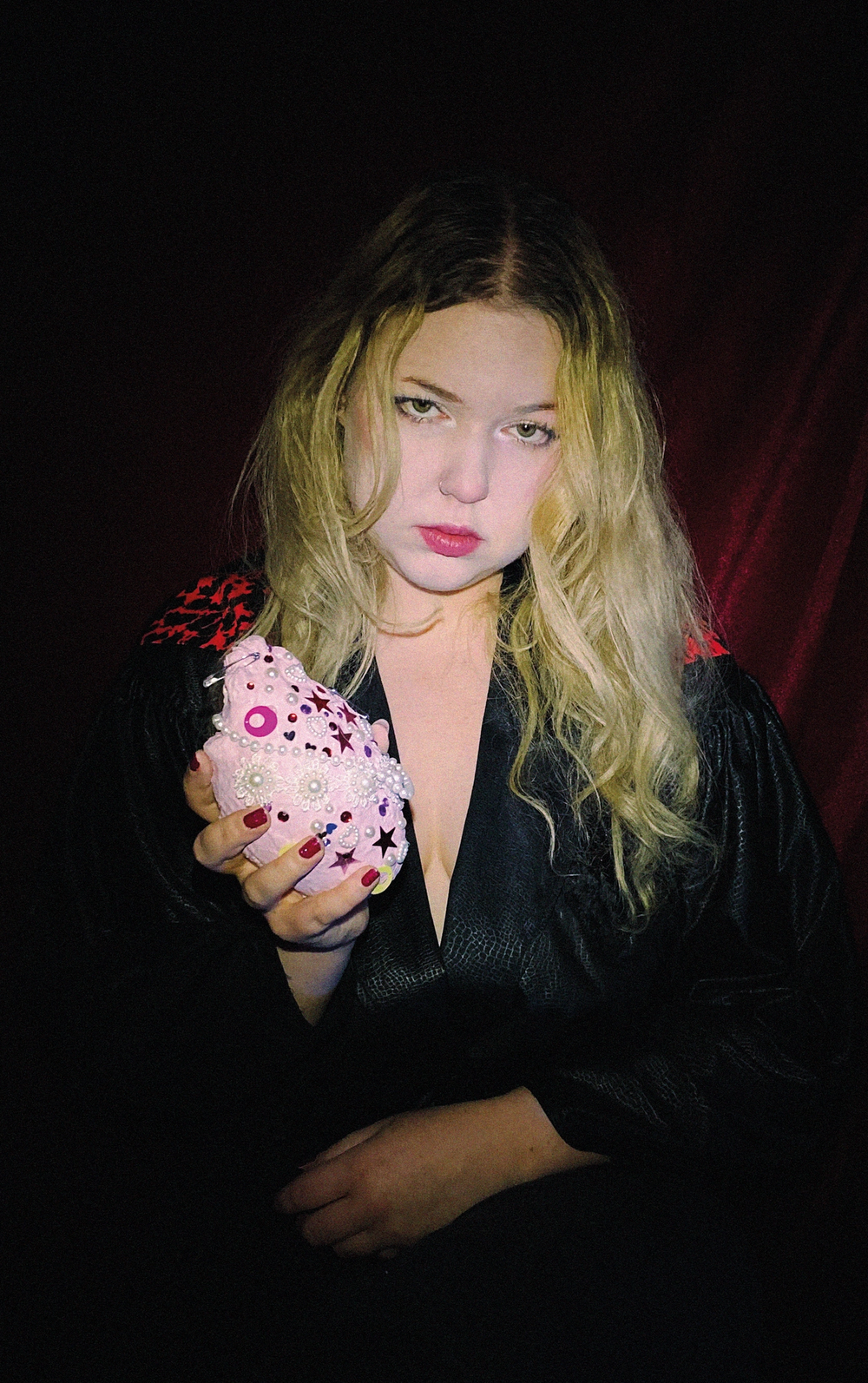 Low-key portrait in a black robe, holding out a soft pink heart studded with pearls, beads and stars